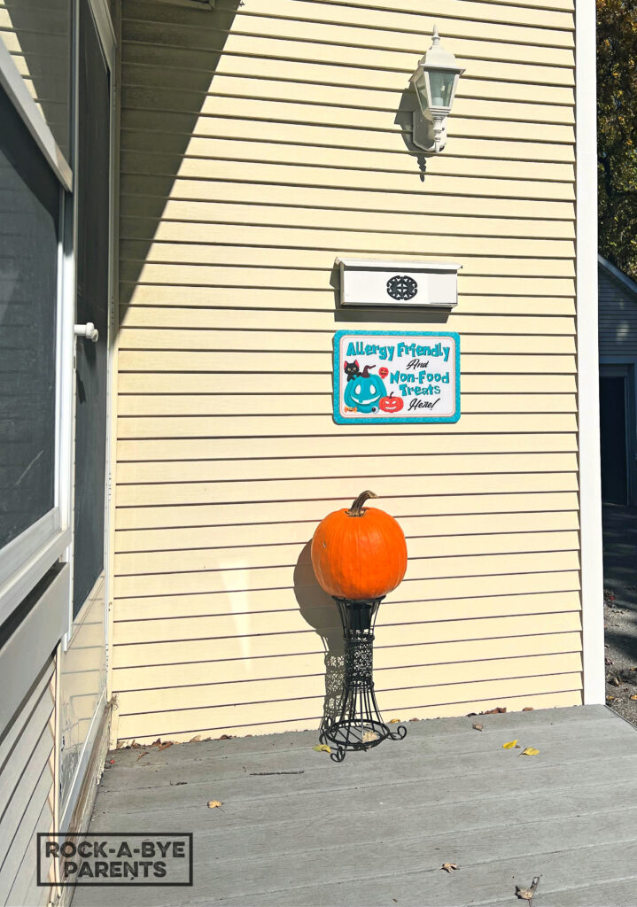A front porch with a mailbox and underneath it is a sign with a black cat on a teal jack-o-lantern and it says "Allergy Friendly and Non-Food Treats Here!" Below the sign is a real pumpkin on a black garden stand.