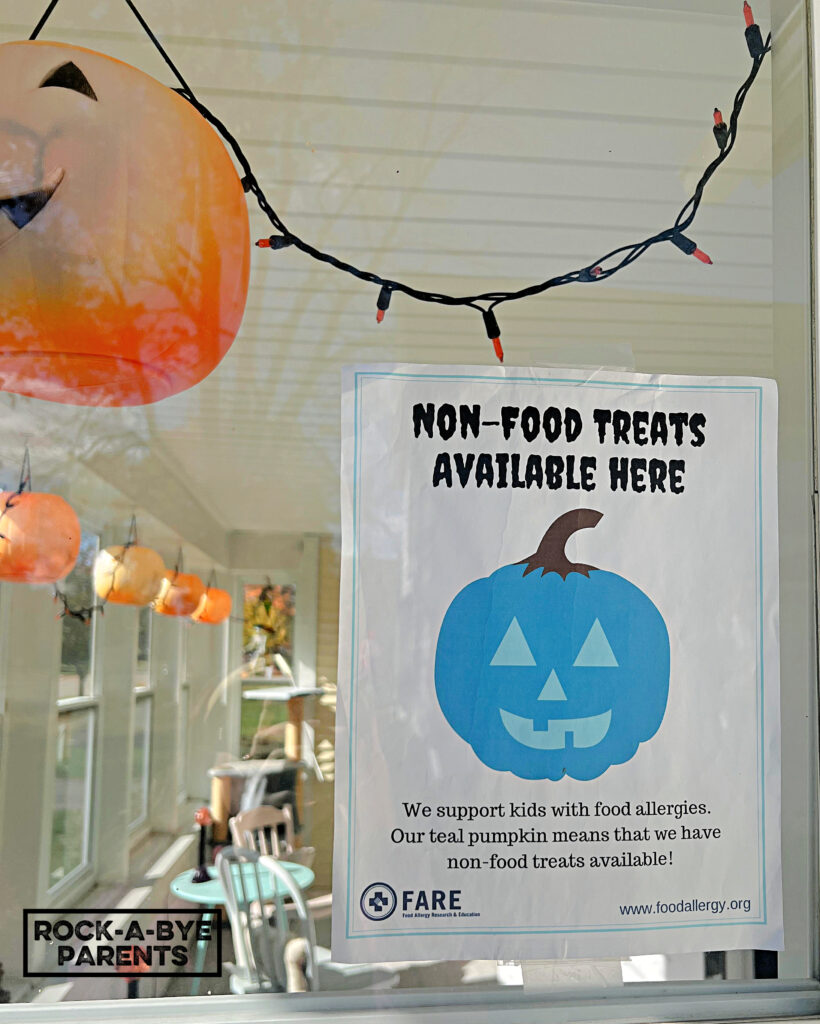 The window of a front porch at Halloween time. The window has a printable sign from FARE that has a teal pumpkin on it and says "Non-Food Treats Available Here."