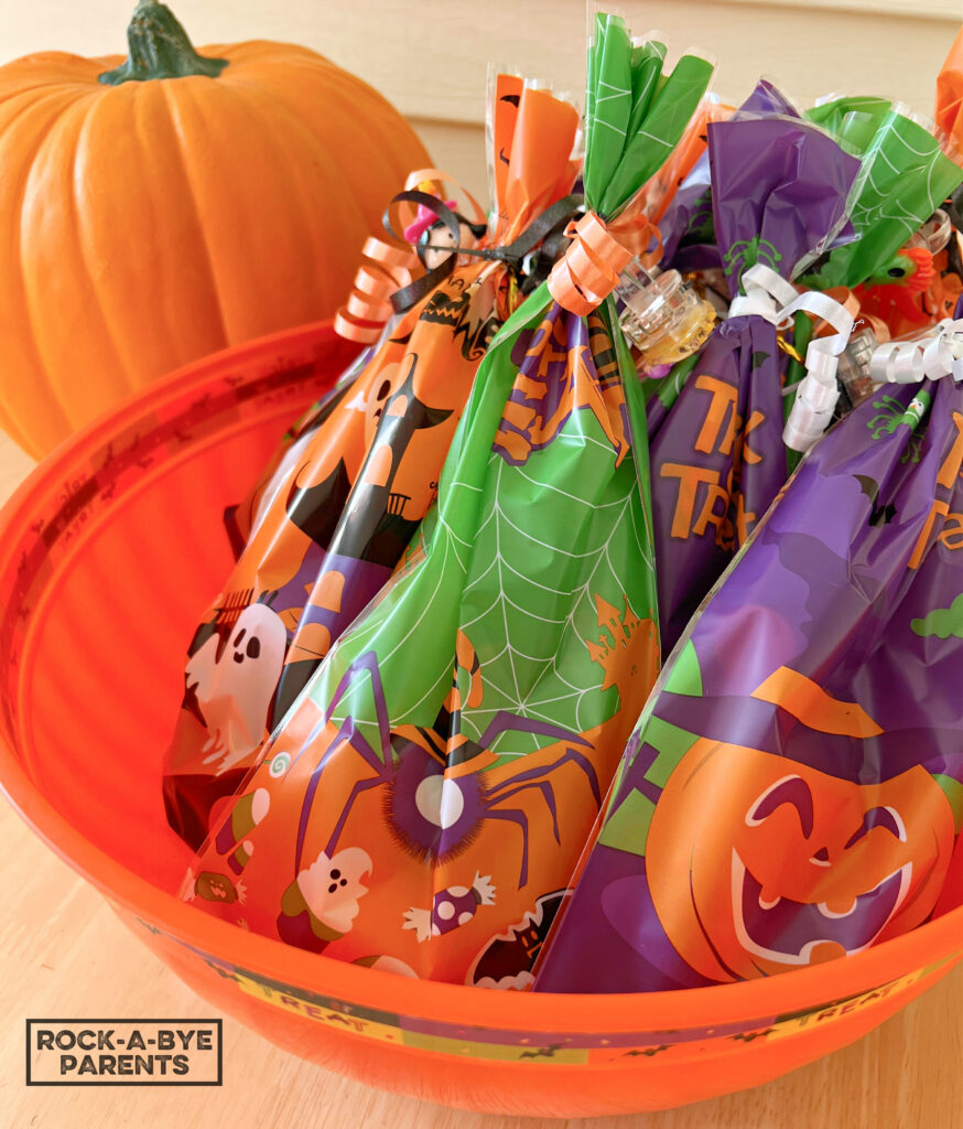 A large orange Halloween bowl full of Halloween non-candy goodie bags in different colors. The bowl is sitting on a light brown table. In the background is a soft, realistic looking, fake pumpkin.