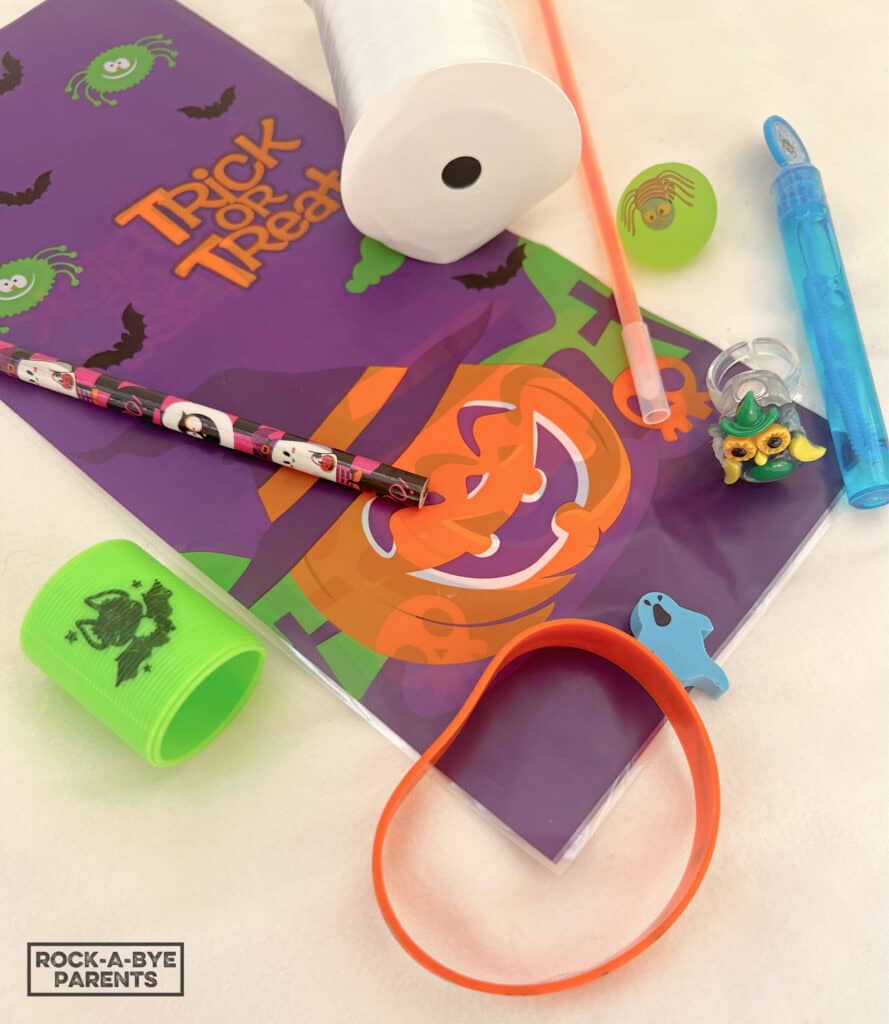 Image of supplies needed to make candy-free Halloween treat bags. Items, lined up on a cream background, are a plastic Halloween themed treat bag, a Halloween pencil, green spring coil with a bat image, orange bracelet, ghost eraser, blue bubble wand with a spider, large clear ring with an owl, green bouncy ball with a spider, orange glow bracelet, and spool of white curling ribbon.