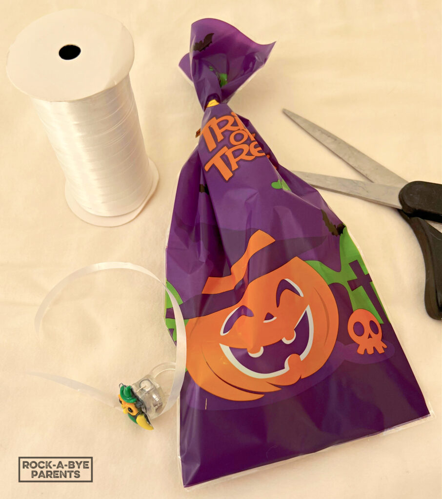 A Halloween treat bag that is closed with a twist tie, a spool of white curling ribbon, a piece of the cut curling ribbon with a Halloween ring, and a pair of scissors.