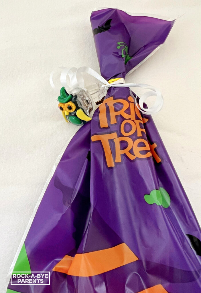 White black ground with a purple Halloween goodie bag tied closed with a white curled ribbon with an owl ring tied onto the ribbon.